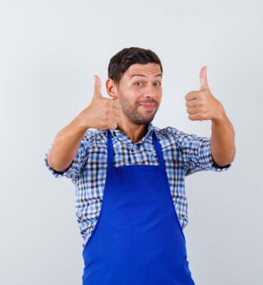 young cook man showing thumbs up in shirt, apron and looking happy , front view.