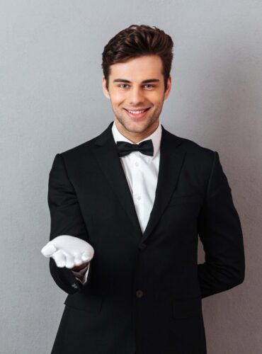 Portrait of a happy smiling man dressed in tuxedo and gloves standing with outsretched hand isolated over gray background